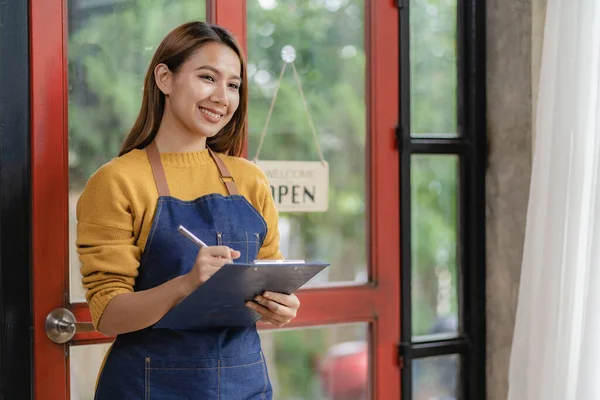 A happy waitress stands at the entrance to the restaurant and looks at the camera. A young businesswoman in an apron stands with an open sign at the entrance with a folder.