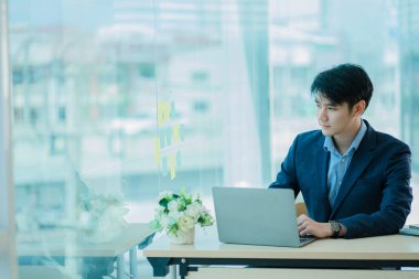 Ofiste başarılı bir laptop kullanan yakışıklı Asyalı işadamı, siyah takım elbiseli ciddi bir işadamı büyük pencerenin yanında dizüstü bilgisayarına bir şeyler yazıyor..