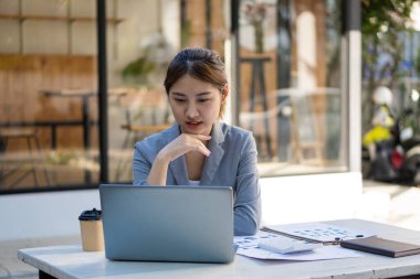 Ahşap masa, vergi, muhasebe, istatistiksel raporlar ve finansal grafikli analitik araştırma kavramları üzerinde matematik finansmanı için hesap makinesi ve dizüstü bilgisayar kullanan Asyalı kadın.