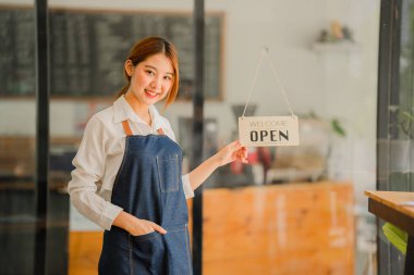 Dükkanının önünde duran genç bir esnaf. Bir dükkan aç. Önlüklü güzel bir Asyalı kadın tablet tutuyor ve açık bir tabelayla bir kafe kapısının önünde duruyor. işletme sahibi başlangıç fikirleri