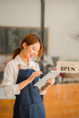 Dükkanının önünde duran genç bir esnaf. Bir dükkan aç. Önlüklü güzel bir Asyalı kadın tablet tutuyor ve açık bir tabelayla bir kafe kapısının önünde duruyor. işletme sahibi başlangıç fikirleri