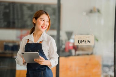 Dükkanının önünde duran genç bir esnaf. Bir dükkan aç. Önlüklü güzel bir Asyalı kadın tablet tutuyor ve açık bir tabelayla bir kafe kapısının önünde duruyor. işletme sahibi başlangıç fikirleri