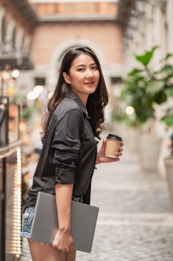 Siyah gömlekli güzel bir Asyalı kadın elinde kahve bardağıyla bir alışveriş merkezinin önünde duruyor..
