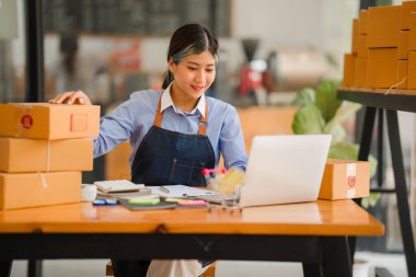 Küçük işletme kuruluşu, KME, gülümseyen genç Asyalı başarılı girişimcilerin sahibi. Paket hazırlamak için akıllı telefon ya da dizüstü bilgisayar kullanınız ve online siparişleri kontrol ediniz. İnternet üzerinden fikir satmak