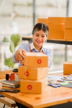 Küçük işletme kuruluşu, KME, gülümseyen genç Asyalı başarılı girişimcilerin sahibi. Paket hazırlamak için akıllı telefon ya da dizüstü bilgisayar kullanınız ve online siparişleri kontrol ediniz. İnternet üzerinden fikir satmak