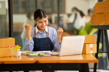 Küçük işletme kuruluşu, KME, gülümseyen genç Asyalı başarılı girişimcilerin sahibi. Paket hazırlamak için akıllı telefon ya da dizüstü bilgisayar kullanınız ve online siparişleri kontrol ediniz. İnternet üzerinden fikir satmak