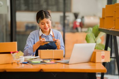 Küçük işletme kuruluşu, KME, gülümseyen genç Asyalı başarılı girişimcilerin sahibi. Paket hazırlamak için akıllı telefon ya da dizüstü bilgisayar kullanınız ve online siparişleri kontrol ediniz. İnternet üzerinden fikir satmak
