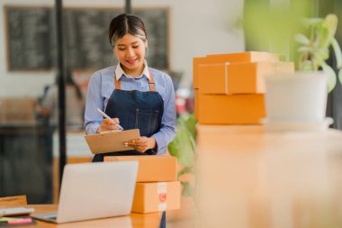 Küçük işletme kuruluşu, KME, gülümseyen genç Asyalı başarılı girişimcilerin sahibi. Paket hazırlamak için akıllı telefon ya da dizüstü bilgisayar kullanınız ve online siparişleri kontrol ediniz. İnternet üzerinden fikir satmak