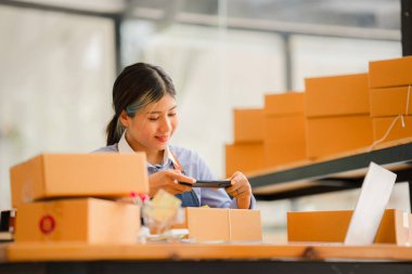 Küçük işletme kuruluşu, KME, gülümseyen genç Asyalı başarılı girişimcilerin sahibi. Paket hazırlamak için akıllı telefon ya da dizüstü bilgisayar kullanınız ve online siparişleri kontrol ediniz. İnternet üzerinden fikir satmak