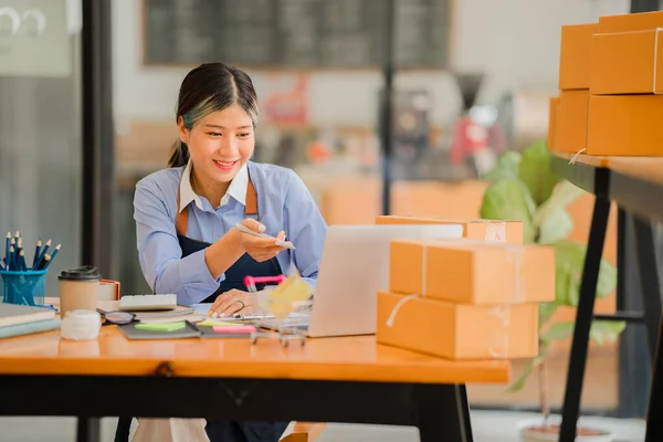 Small business start-up, SME, owner of successful young Asian woman entrepreneurs smiling. use a smartphone or laptop Receive and check online orders to prepare packing boxes. selling ideas online