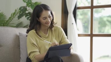 Mutlu Asyalı kadın evinde oturmuş elinde dijital tabletle internet sörfü çalışmalarının keyfini çıkarıyor.