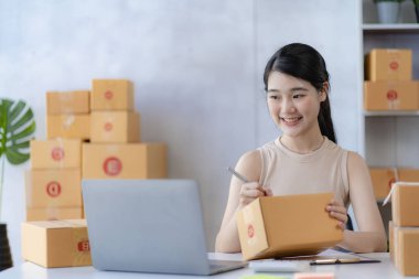 İş dünyasından tatlı Asyalı kadın evde dizüstü bilgisayarla satış yapıyor siparişleri alıyor ve sarı paketleri çevrimiçi dağıtıyor.