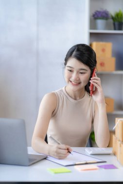 İş dünyasından tatlı Asyalı kadın evde dizüstü bilgisayarla satış yapıyor siparişleri alıyor ve sarı paketleri çevrimiçi dağıtıyor.