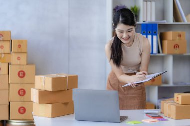 İş dünyasından tatlı Asyalı kadın evde dizüstü bilgisayarla satış yapıyor siparişleri alıyor ve sarı paketleri çevrimiçi dağıtıyor.