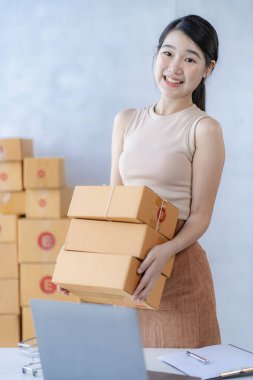 İş dünyasından tatlı Asyalı kadın evde dizüstü bilgisayarla satış yapıyor siparişleri alıyor ve sarı paketleri çevrimiçi dağıtıyor.