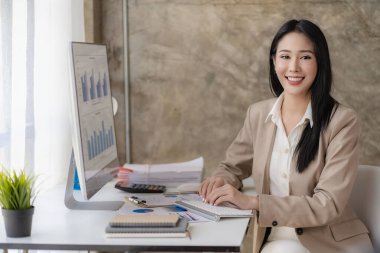 Bilgisayarda çalışan Asyalı iş kadını, hesap makinesi ve iş raporuyla birlikte modern pencere kenarındaki ofiste finansal grafik belgesi ile birlikte çalışıyor.