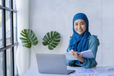 Güzel bir Asyalı Müslüman kadın başörtüsü takıyor. Evindeki oturma odasında finansal muhasebe işlerini yapmak için bilgisayarını kullanıyor..