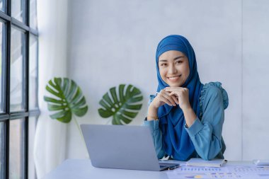 Güzel bir Asyalı Müslüman kadın başörtüsü takıyor. Evindeki oturma odasında finansal muhasebe işlerini yapmak için bilgisayarını kullanıyor..