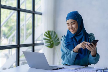 Güzel bir Asyalı Müslüman kadın akıllı telefon ve dizüstü bilgisayarla evindeki oturma odasında finansal muhasebe işi yapmak için başörtüsü takıyor..