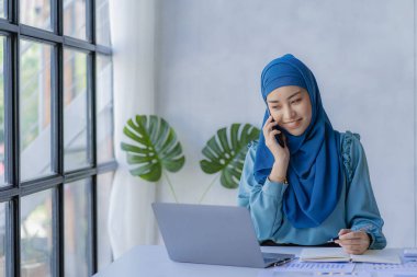 Güzel bir Asyalı Müslüman kadın akıllı telefon ve dizüstü bilgisayarla evindeki oturma odasında finansal muhasebe işi yapmak için başörtüsü takıyor..