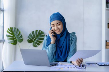 Güzel bir Asyalı Müslüman kadın akıllı telefon ve dizüstü bilgisayarla evindeki oturma odasında finansal muhasebe işi yapmak için başörtüsü takıyor..