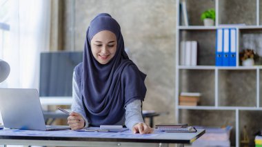 Güzel bir Asyalı Müslüman kadın başörtüsü takıyor. Evindeki oturma odasında finansal muhasebe işlerini yapmak için bilgisayarını kullanıyor..
