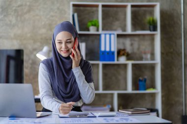 Güzel bir Asyalı Müslüman kadın akıllı telefon ve dizüstü bilgisayarla evindeki oturma odasında finansal muhasebe işi yapmak için başörtüsü takıyor..