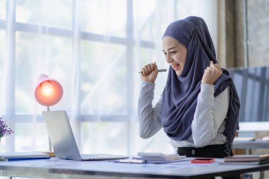 Güzel bir Asyalı Müslüman kadın başörtüsü takıyor. Evindeki oturma odasında finansal muhasebe işlerini yapmak için bilgisayarını kullanıyor..