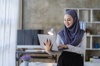 Güzel bir Asyalı Müslüman kadın başörtüsü takıyor. Evindeki oturma odasında finansal muhasebe işlerini yapmak için bilgisayarını kullanıyor..