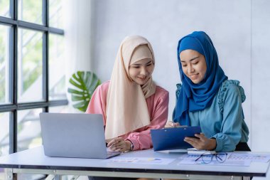 Ofiste finansal grafiklerle çalışan iki güzel Asyalı Müslüman kadın. Geleneksel giysili Müslüman iş kadını ofis toplantısında çalışıyor ve konuşuyor.