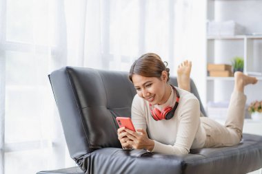 Kanepede oturan Asyalı kadın akıllı telefonlu kulaklık takarak film izleyip evde müzik dinliyor.