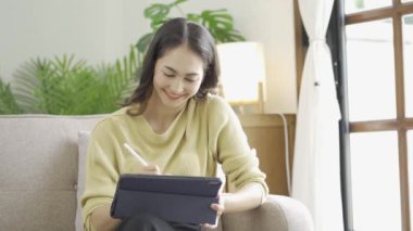 Rahat bir koltukta oturan hoş bir Asyalı kadın. evde bir tablet bilgisayar kullanın mutlu kız veri tarıyor popüler online mağazalarda alışveriş yapıyor e-kitap okuyor