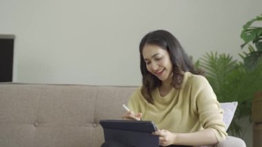 Rahat bir koltukta oturan hoş bir Asyalı kadın. evde bir tablet bilgisayar kullanın mutlu kız veri tarıyor popüler online mağazalarda alışveriş yapıyor e-kitap okuyor