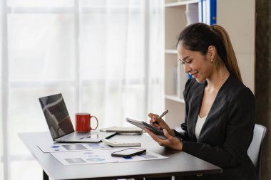 İş yerinde oturan Asyalı iş kadını, analitik istatistikleri hesaplamak için hesap makinesi kullanıyor. Elinde kalem ve grafik kağıdı olan güzel bir kadının ofisteki bilgisayarını görüyor.