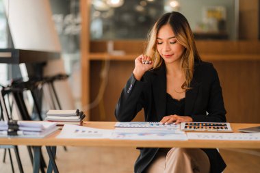 İş yerinde oturan Asyalı iş kadını, analitik istatistikleri hesaplamak için hesap makinesi kullanıyor. Elinde kalem ve grafik kağıdı olan güzel bir kadının ofisteki bilgisayarını görüyor.