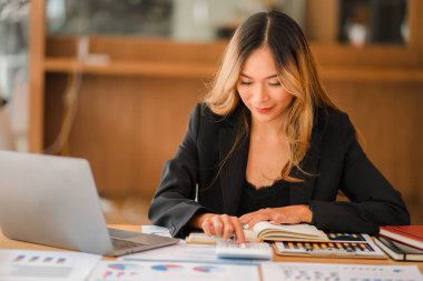 İş yerinde oturan Asyalı iş kadını, analitik istatistikleri hesaplamak için hesap makinesi kullanıyor. Elinde kalem ve grafik kağıdı olan güzel bir kadının ofisteki bilgisayarını görüyor.