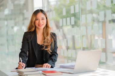 İş yerinde oturan Asyalı iş kadını, analitik istatistikleri hesaplamak için hesap makinesi kullanıyor. Elinde kalem ve grafik kağıdı olan güzel bir kadının ofisteki bilgisayarını görüyor.