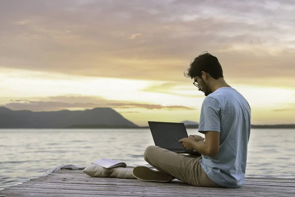 young digital nomad man sitting on wooden pier at sea working on internet remotely at sunset - Traveling with a computer - Online dream job concep