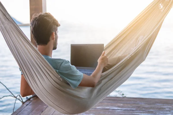 young digital nomad lying on hammock with sea views working remotely on internet at sunset - Traveling with a computer - Dream online job concept - Selective focu