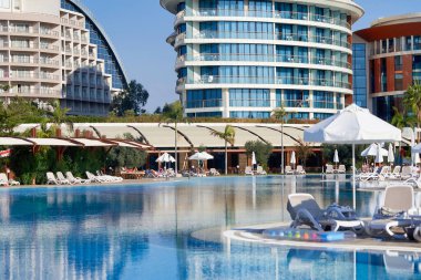 Mavi suyu olan bir havuz, otelin arka planında güneşlik ve şemsiyeler. Otelde turistler için dinlenme yeri
