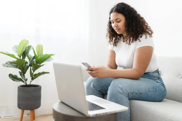 Attraktiv ung afrikansk kvinna sitter på soffan hemma med mobiltelefon och bärbar dator — Stockfoto