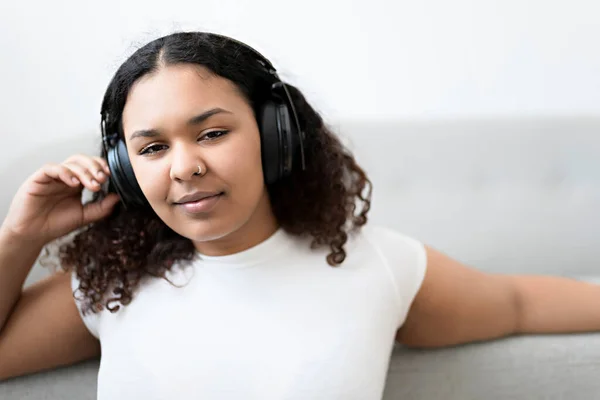 Attractive young African woman sitting on the couch at home listening music with headphone — Stock Photo, Image