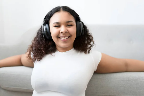 Attractive young African woman sitting on the couch at home listening music with headphone — Stock Photo, Image