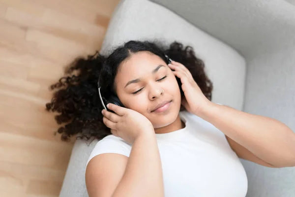 Jovem mulher africana atraente deitada no sofá em casa ouvindo música com fone de ouvido — Fotografia de Stock