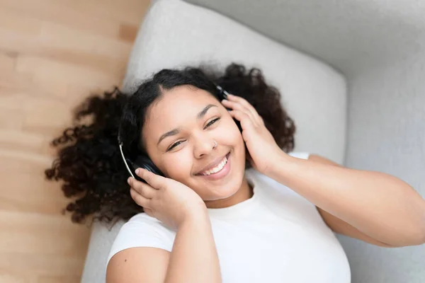 Attractive young African woman lay on the couch at home listening music with headphone — Stock Photo, Image