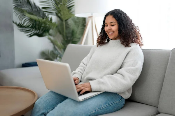 Ελκυστική νεαρή Αφρικανή που εργάζεται σε φορητό υπολογιστή, ενώ κάθεται στον καναπέ στο σπίτι — Φωτογραφία Αρχείου