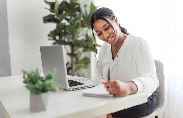 Νεαρή Αφροαμερικανή επιχειρηματίας που εργάζεται στο φορητό υπολογιστή στο γραφείο στο σπίτι με στυλό και σημειωματάριο — Φωτογραφία Αρχείου