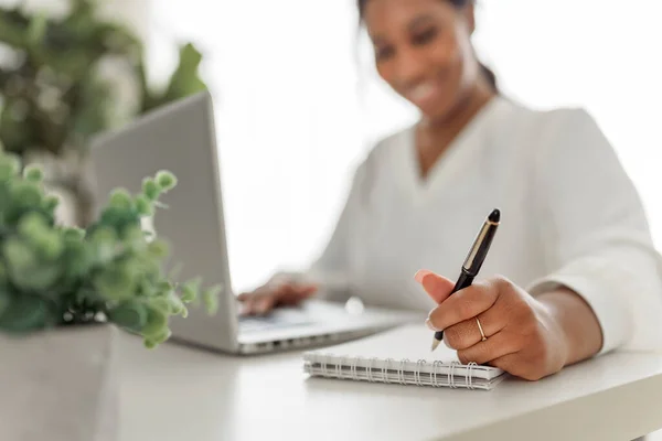 Νεαρή Αφροαμερικανή επιχειρηματίας που εργάζεται στο φορητό υπολογιστή στο γραφείο στο σπίτι με στυλό και σημειωματάριο — Φωτογραφία Αρχείου