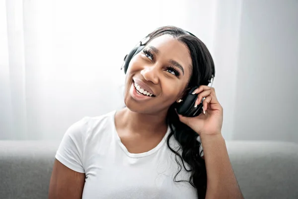 Attractive young African woman sitting on the couch at home listening music with headphone — Stock Photo, Image