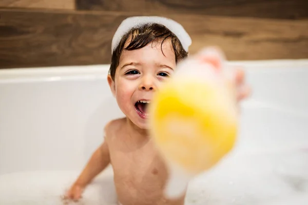 Happy two year kid bathes with toys in bath with foam and duck, — Photo